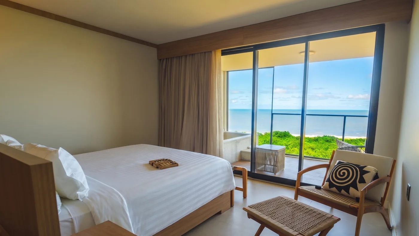 Apartamento frente mar com cama de casal, poltronas e porta de vidro aberta para a varanda com vista para o mar