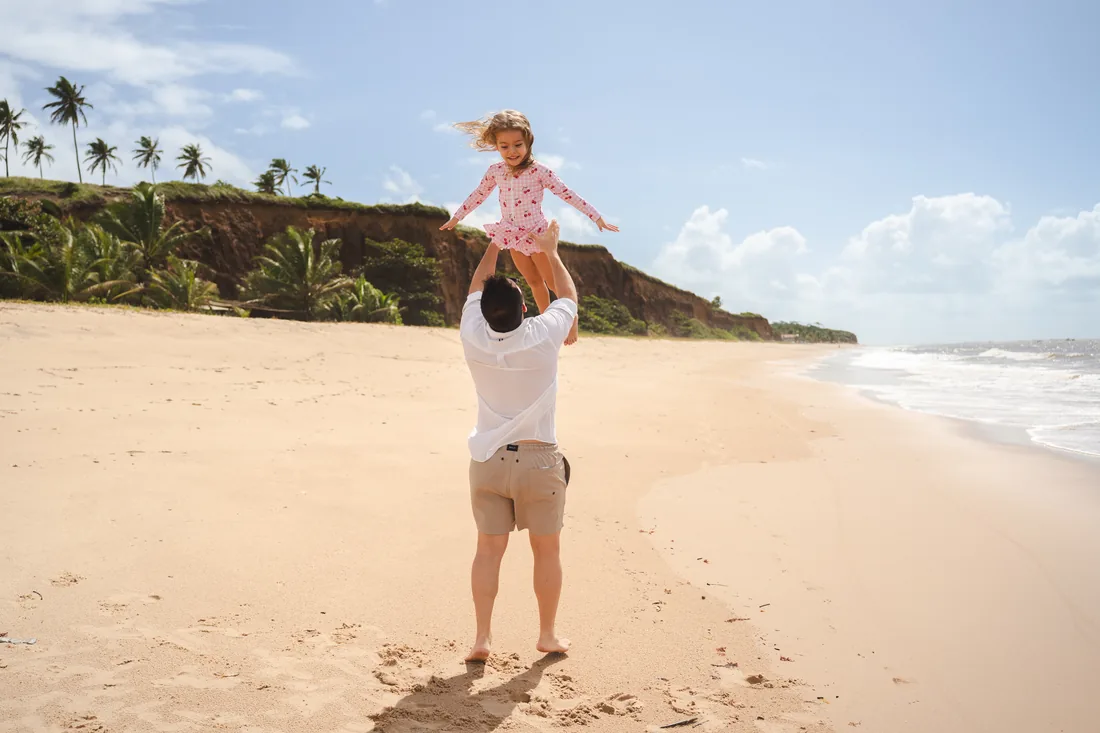 Pai carregando a filha nos ombros na faixa de areia, em frente ao resort