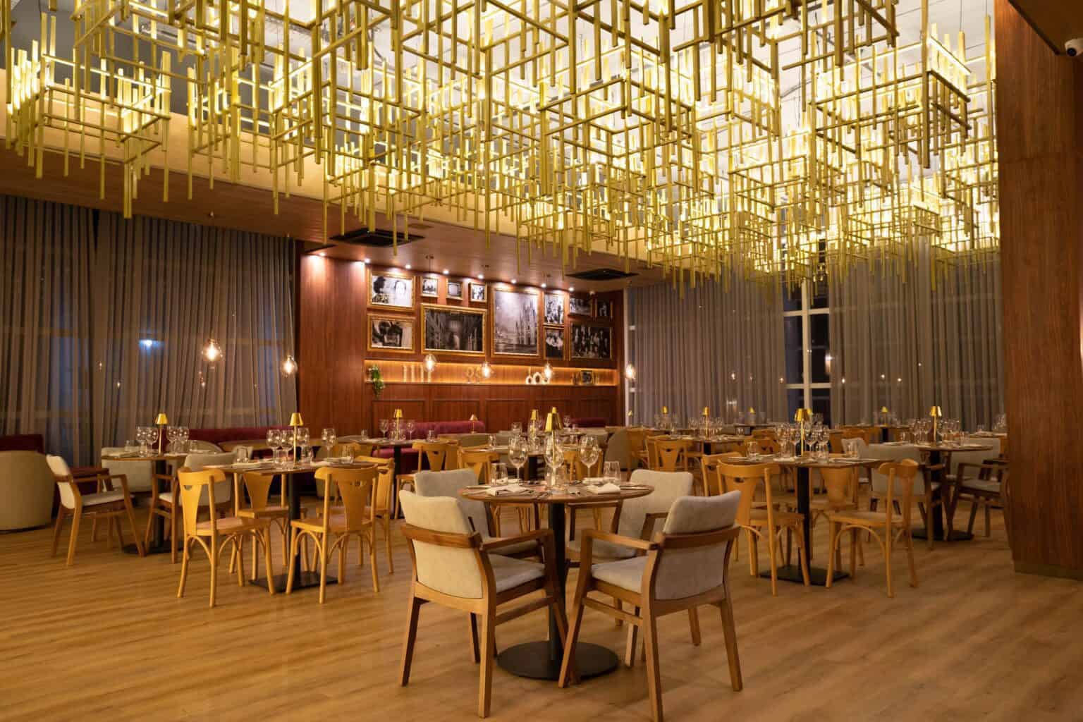 restaurante italiano exclusivo do Tauá ornamentado com luminárias contemporâneas douradas, quadros em preto e branco na parede e mesas postas em espaço amplo com grandes janelas.