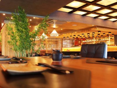 restaurante japonês decorado com painéis ilustrados com gravuras típicas e luminárias no estilo tradicional nipônico. Mesa posta com cerâmicas escuras e arborização com ramos de bambu à esquerda.