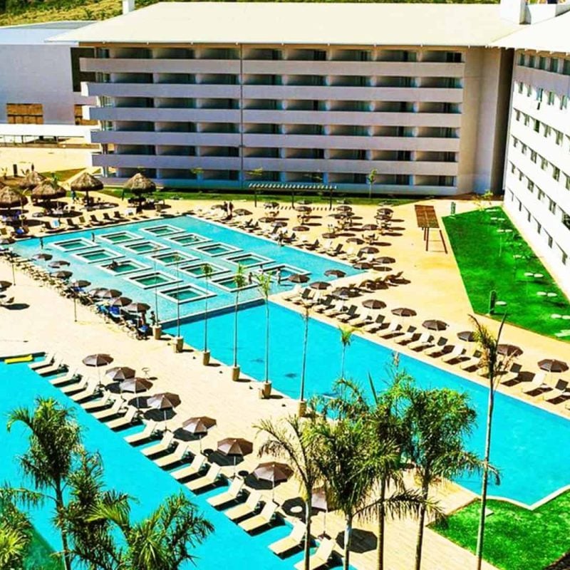 Vista do complexo de piscinas, com jacuzzis ao fundo da imagem, espreguiçadeiras à beira da piscina, ambiente arborizado com palmeiras.