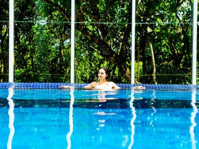 Mulher aprecia um banho de piscina com vista para muita natureza em fundo protegido por parede de vidro.