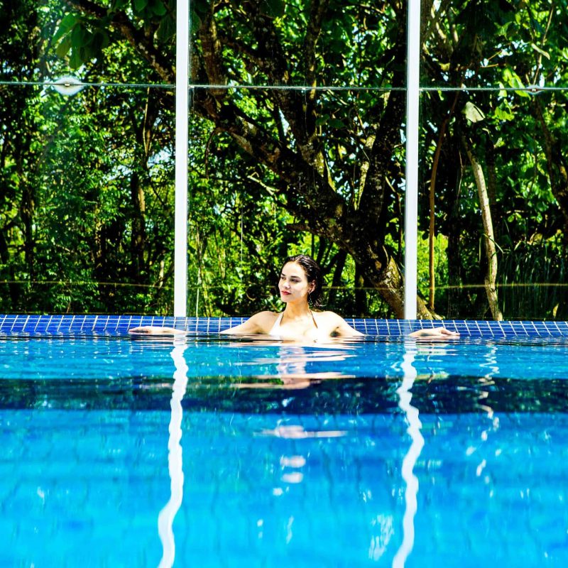 Mulher aprecia um banho de piscina com vista para muita natureza em fundo protegido por parede de vidro.